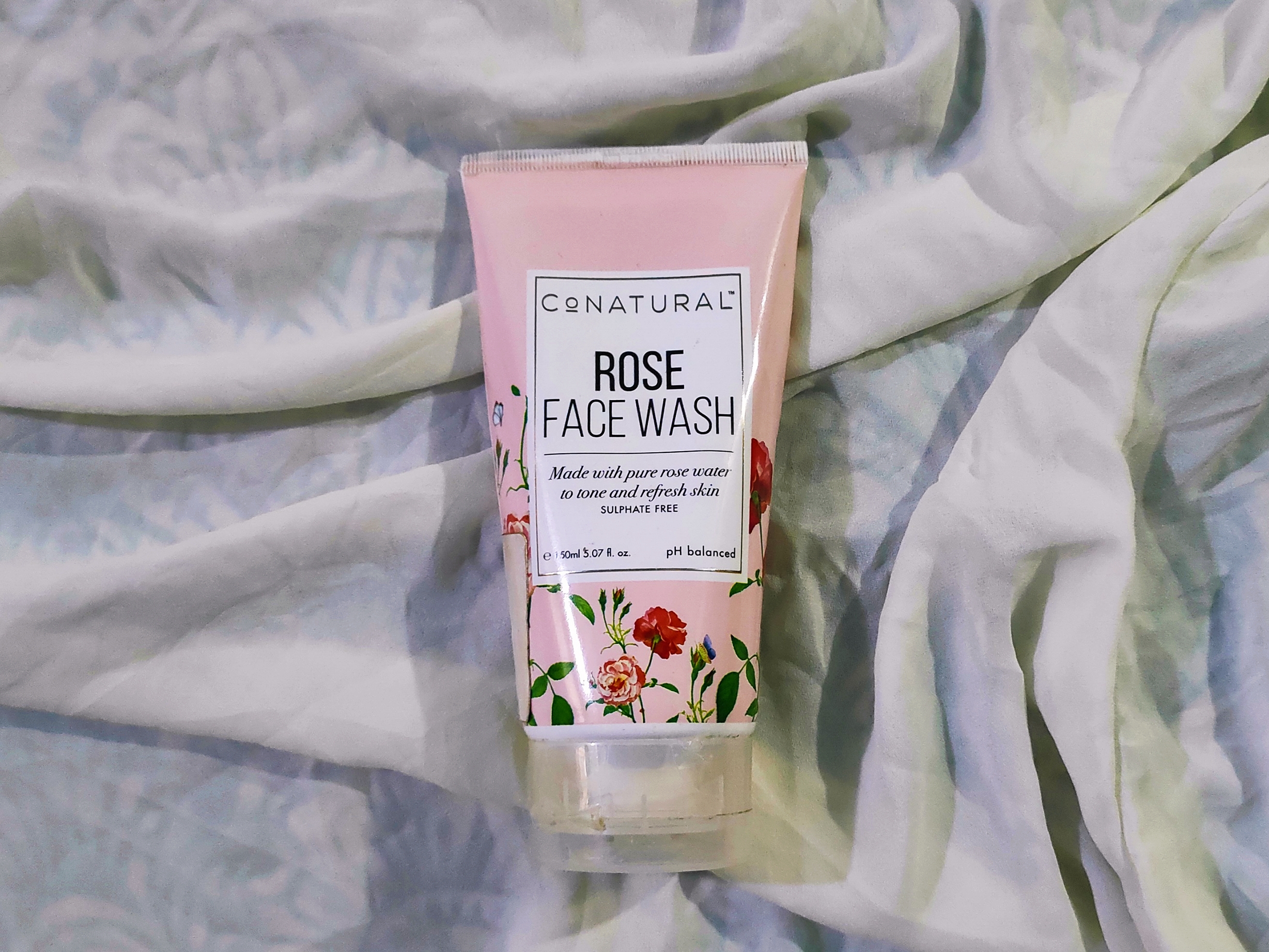 A light pink tube of 150 ml CoNatural rose face wash with a black lable and transparent cap, laying on a white surface.